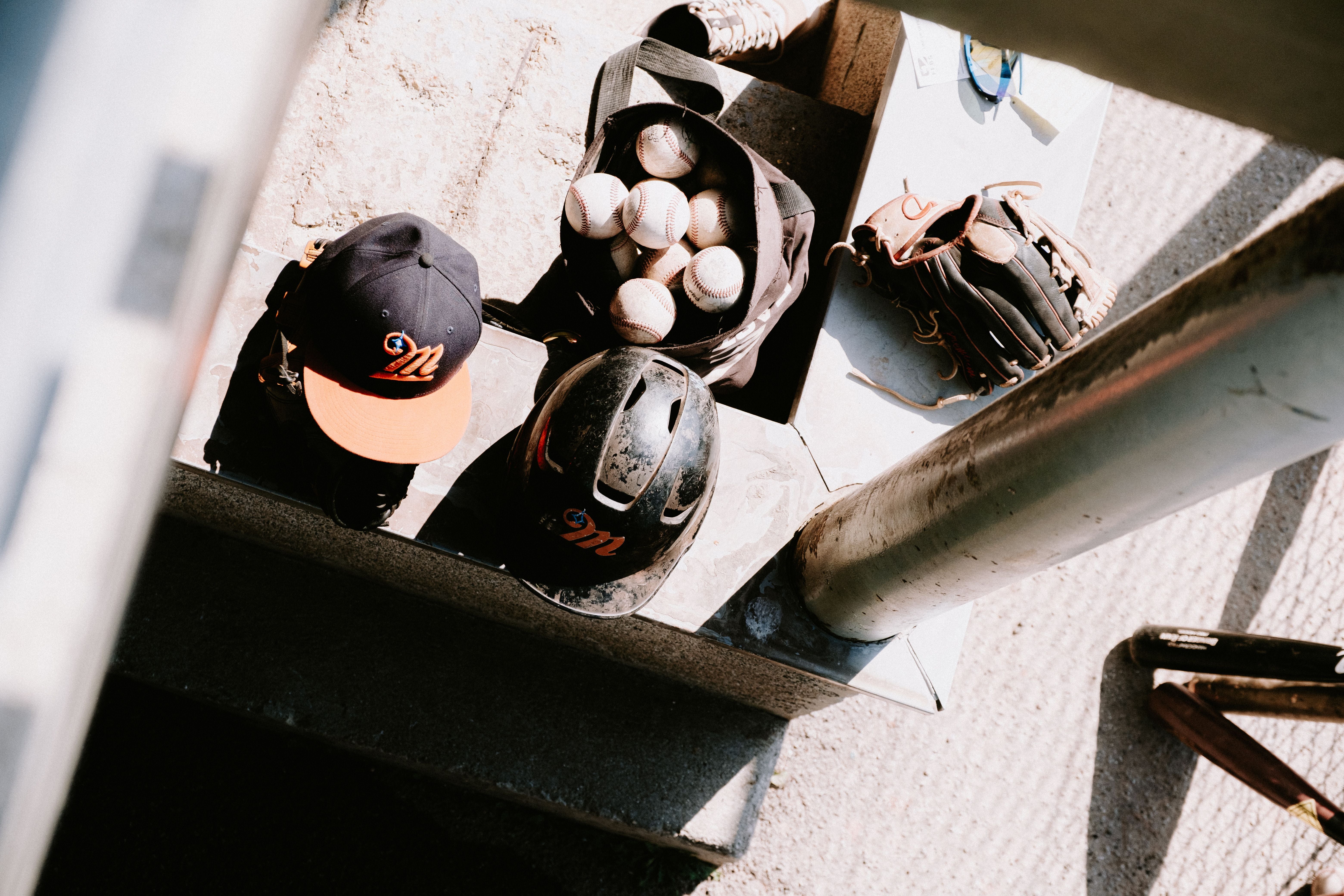 Baseball gear laid out on dugout steps in warm afternoon light — cap, helmet, glove, bat, and a bag of balls